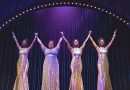 Four black female singers in shimmering white dresses are holding hands raised up in the air. The back of the set is purple with a slight arch of white lights framing the top of the image