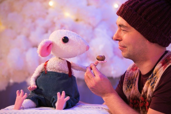Snow Mouse at the egg theatre bath. A man wearing a red hat and bright waistcoat is playing with a small white mouse wearing blue dungarees. He is offering an acorn to the mouse