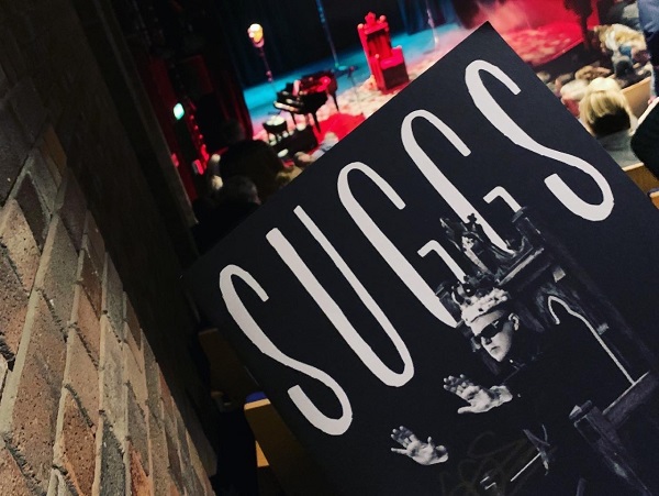 Photograph of a programme for Suggs. The background of the image is slightly blurred but features the stage in the distance with a large red throne surrounded by theatre lights. On the left is the brick wall of the auditorium .