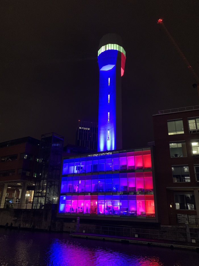 Bristol Light Festival 2022 The Shot Tower in Bristol is lit up in blue and pink lights. The building undernearth is also fully lit with blinds that are going up and down