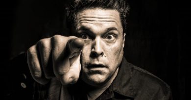 Dom Joly Barn Theatre a black and white photo of Dom Joly with a slight sepia tinge. He's centre frame staring at the camera with a slightly wild look, pointing his finger into the lense at the viewer