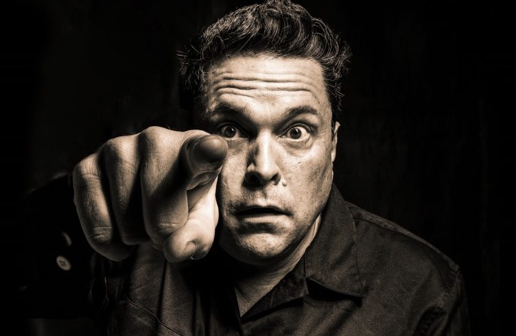 Dom Joly Barn Theatre a black and white photo of Dom Joly with a slight sepia tinge. He's centre frame staring at the camera with a slightly wild look, pointing his finger into the lense at the viewer