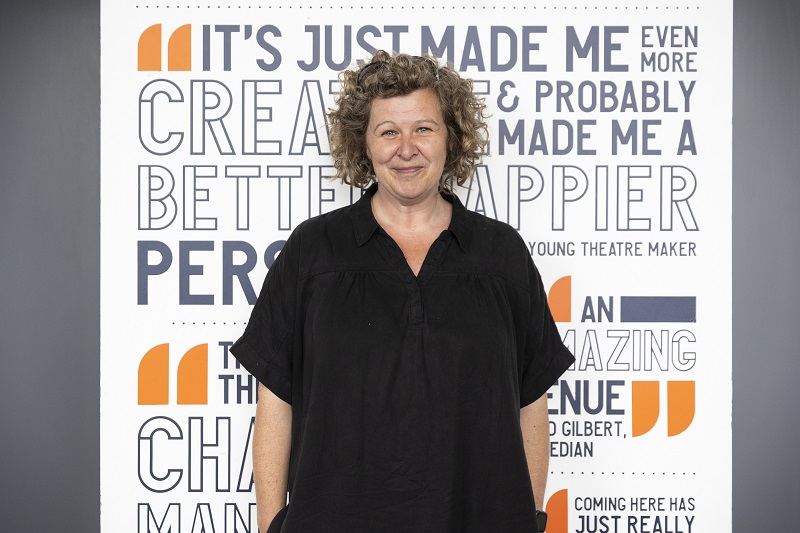 Heidi Vaughan Tobacco Factory Theatres Photo Craig Fuller - Heidi stands facing the camera with a smile. She has short dark curly hair that is jaw length and wears a long black top