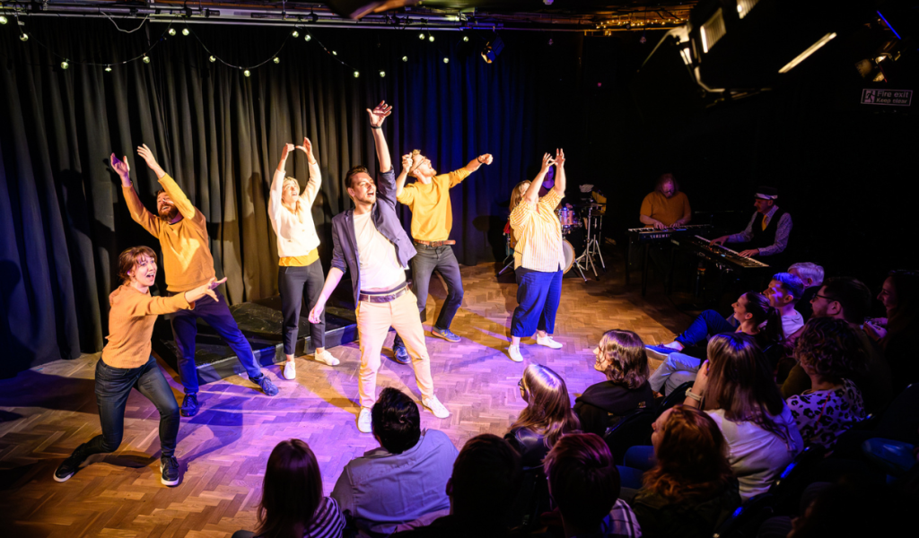 This is Your Musical Bristol Improv Theatre Campaign. A group of actors on stage raise their arms in the air, positioned as if they have finished a finale song