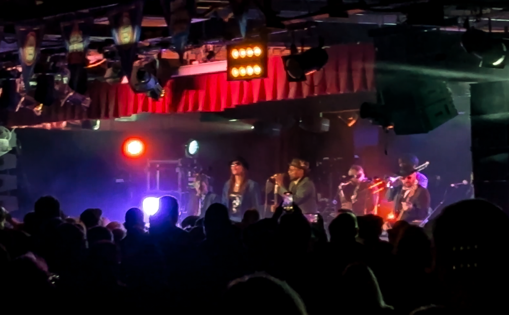 Neville Staple Live Minehead - The Specials former singer is stood on stage with his band. The lighting is all red and purple with the silhouette of a crowd of people in front of the stage